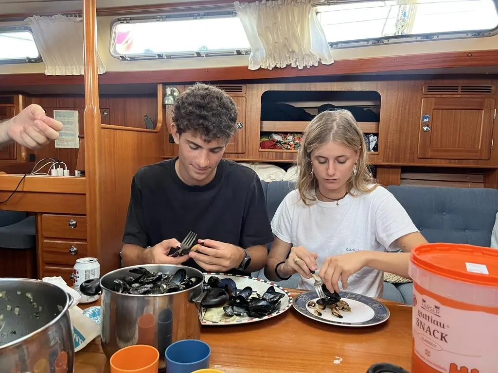 Due persone mangiano cozze sedute al tavolo di una cabina in legno, piatti spaiati e una latta di snack sul tavolo.