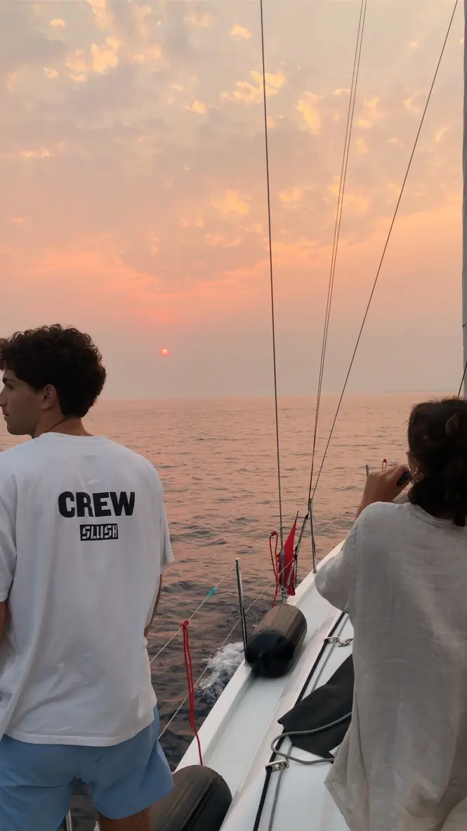 Due persone di spalle a prua guardano il sole calare sul mare, il cielo è rosa; una indossa una maglietta con la scritta CREW.