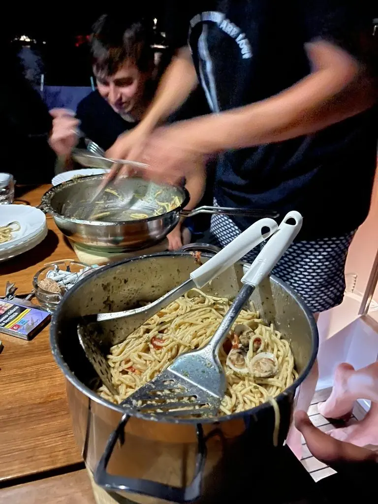 Spaghetti alle vongole serviti da una pentola sul tavolo di bordo, di sera; il braccio di chi serve è mosso.