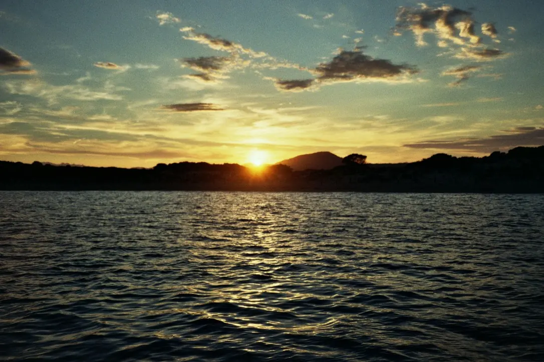 Sole che tocca il profilo delle colline oltre una baia increspata, nuvole lunghe illuminate d’arancio.