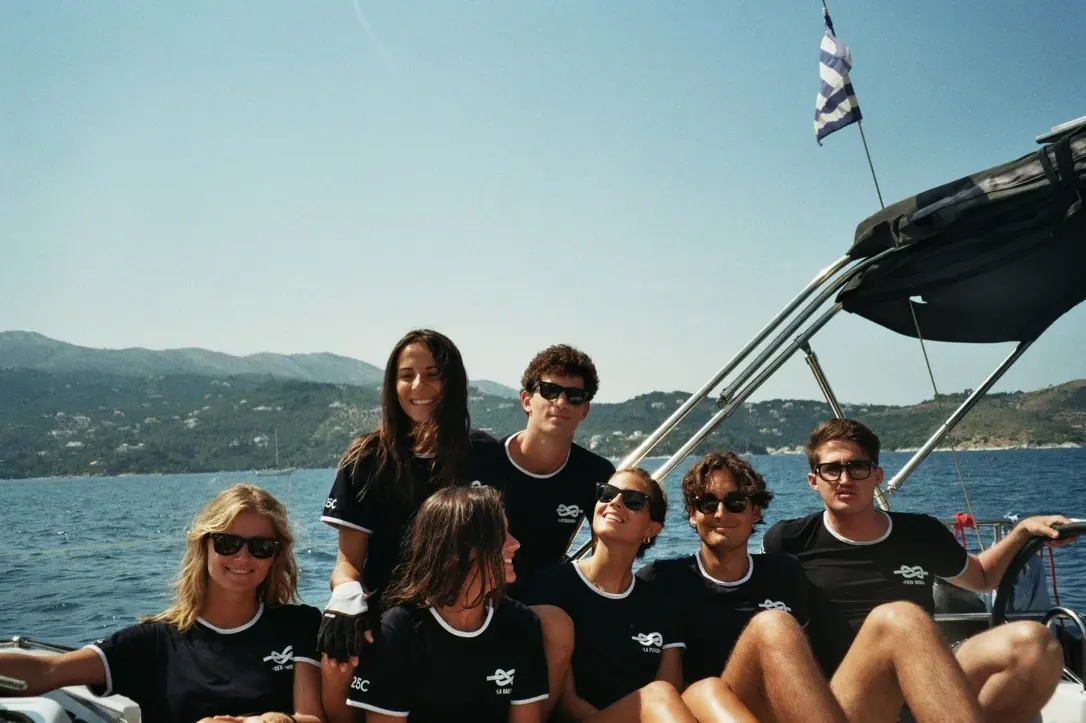 Sette ragazzi in maglietta blu uguale seduti a poppa sotto il bimini, mare e costa verde alle spalle, bandiera greca sull’asta.