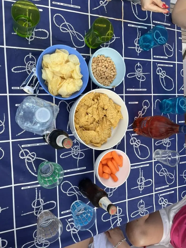 Tavola vista dall’alto su una tovaglia stampata a nodi marinari: ciotole di patatine, arachidi, nachos e carote, bottiglie di birra e aranciata, bicchieri di plastica colorati.