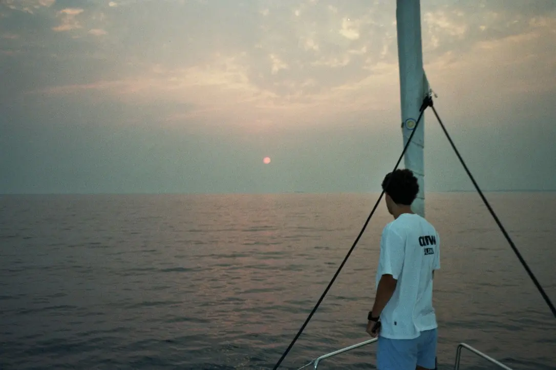 Una persona di spalle a prua guarda il sole basso nella foschia, appoggiata a una sartia; il mare è liscio e il cielo lattiginoso.