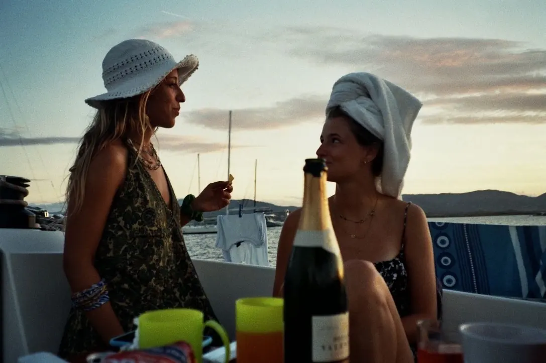 Due ragazze sedute a poppa al tramonto parlano tra loro, una con un asciugamano in testa; sul tavolo una bottiglia e bicchieri di plastica fluo.
