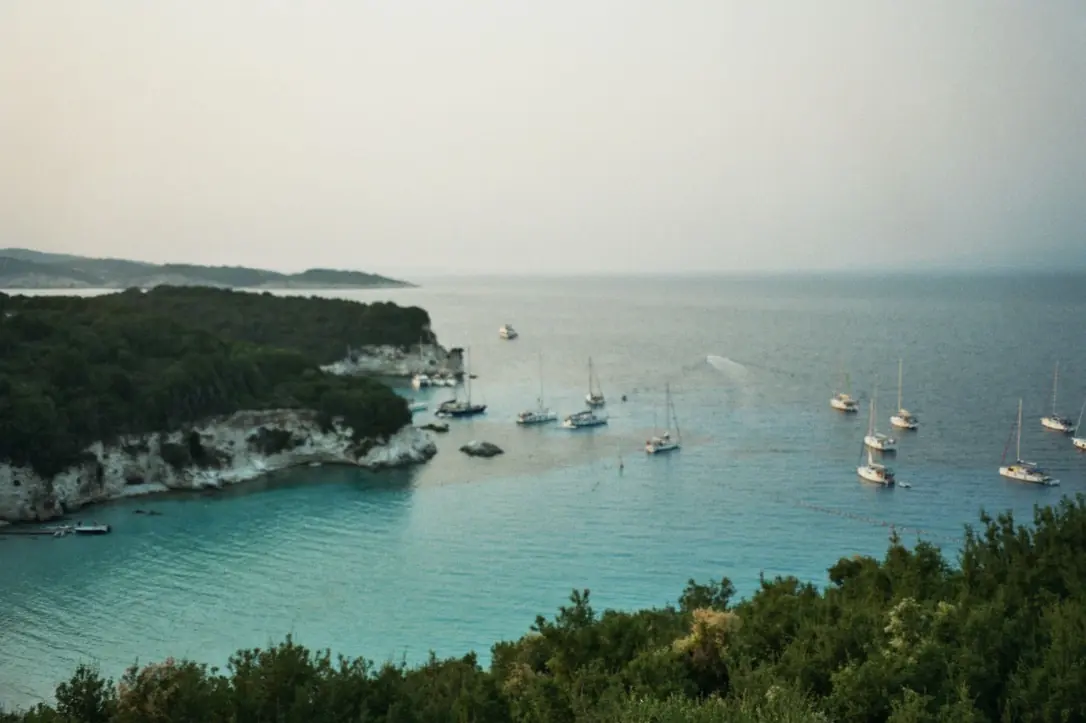 Baia vista dall’alto nella foschia: acqua turchese, una decina di barche a vela alla fonda, scogliera bianca e macchia verde in primo piano.