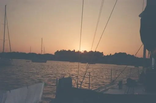 Tramonto visto dal ponte di una barca: sartie in controluce, alberi di altre barche all’ancora, sole basso sull’isola.
